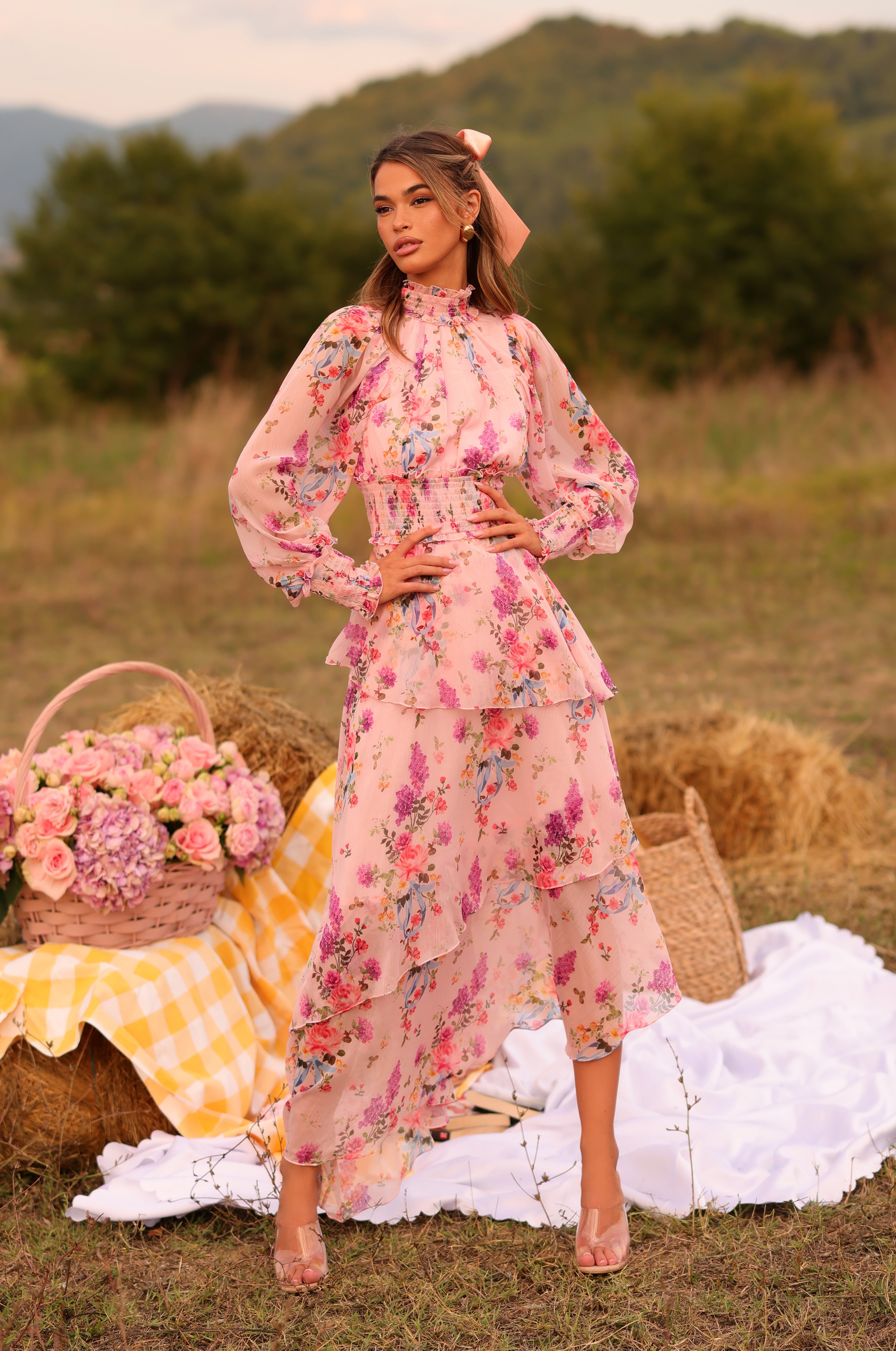 A woman wears the Long Sleeve Blush Pink Floral Maxi Dress: Bellaria while standing on grass near hay bales, a picnic basket, and flowers, surrounded by green trees and hills in the background.